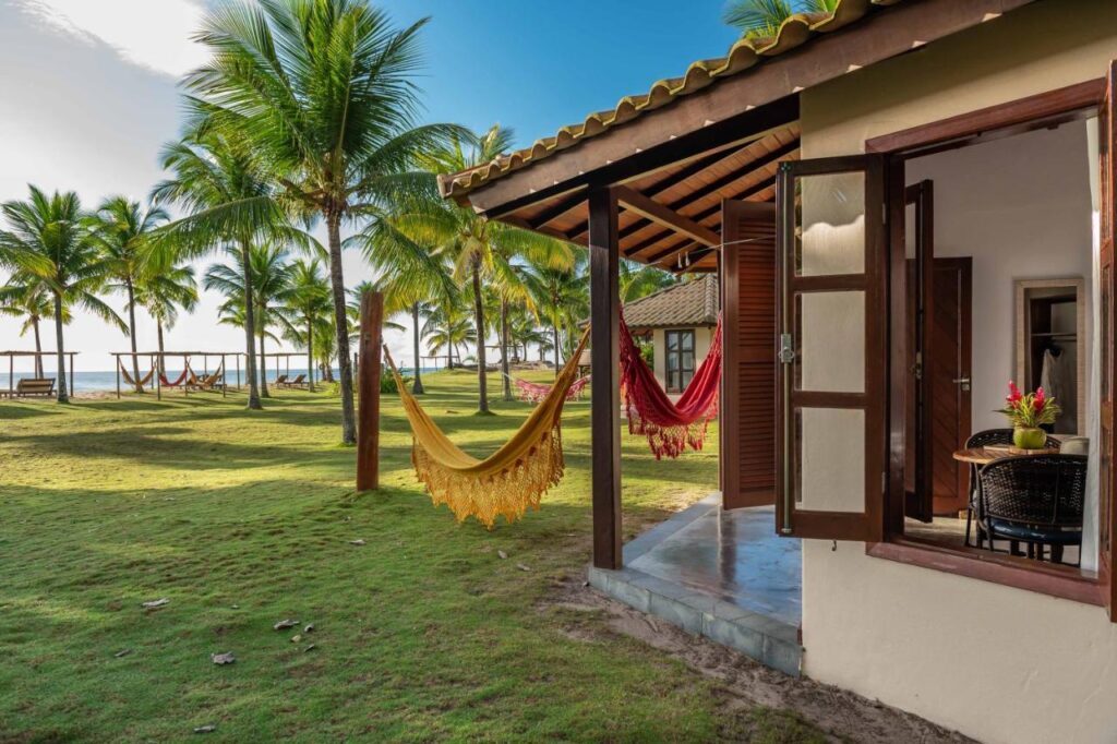 lateral de quarto de hotel com redes em varanda com grama e palmeiras com praia ao fundo