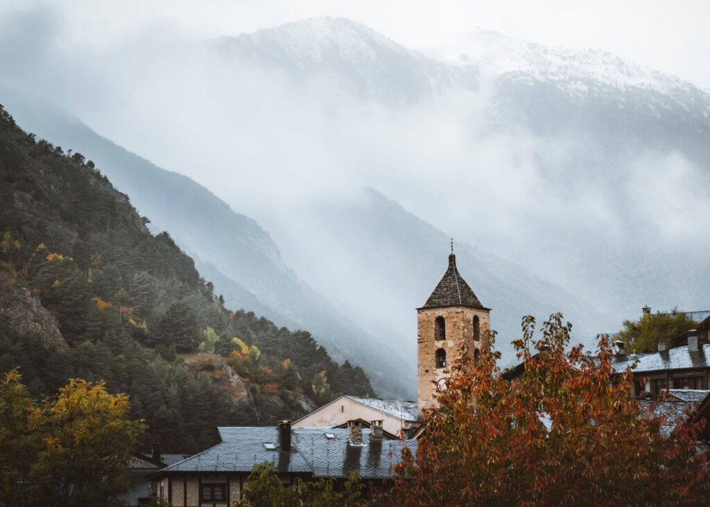 torre de igreja com árvore de outono e montanhas entre nuvens ao fundo